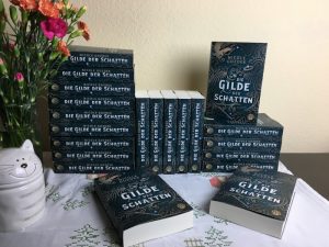 mehrere Bücherstapel von "Die Gilde der Schatten" neben einem Strauß Blumen und einer Dose mit Katzengesicht